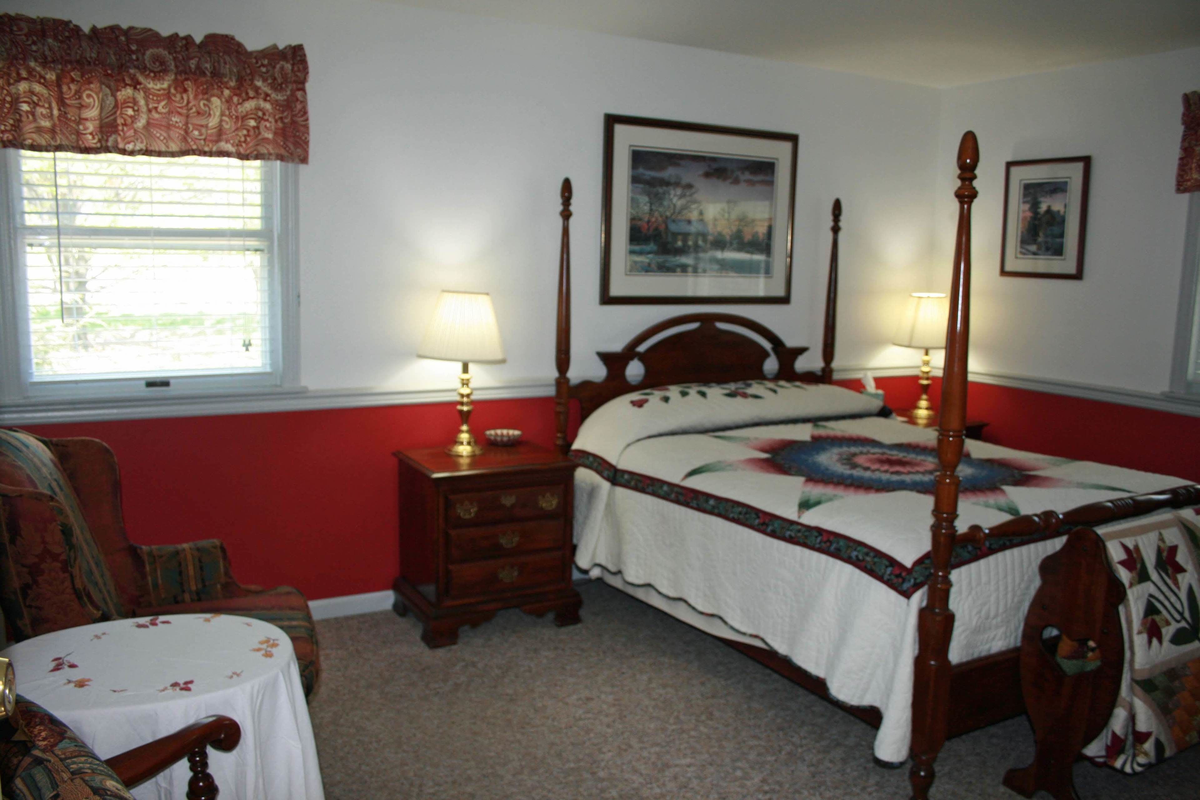 A bedroom with a four-poster bed, two bedside tables with lamps, a chair, and a small table with a floral tablecloth.