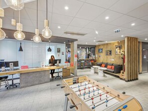 A modern lobby with a reception desk, computers, a foosball table, and a seating area with a guitar. - ibis Saint Germain en Laye Centre (Saint-Germain-en-Laye)