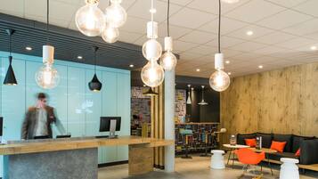 A modern reception area with a wooden counter, hanging pendant lights, and a seating area with orange chairs and a white table.