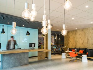 A modern reception area with a wooden counter, hanging pendant lights, and a seating area with orange chairs and a white table. - ibis Saint Germain en Laye Centre (Saint-Germain-en-Laye)