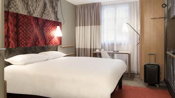 A hotel room with a large bed, a desk, a chair, a suitcase, and a window with curtains.