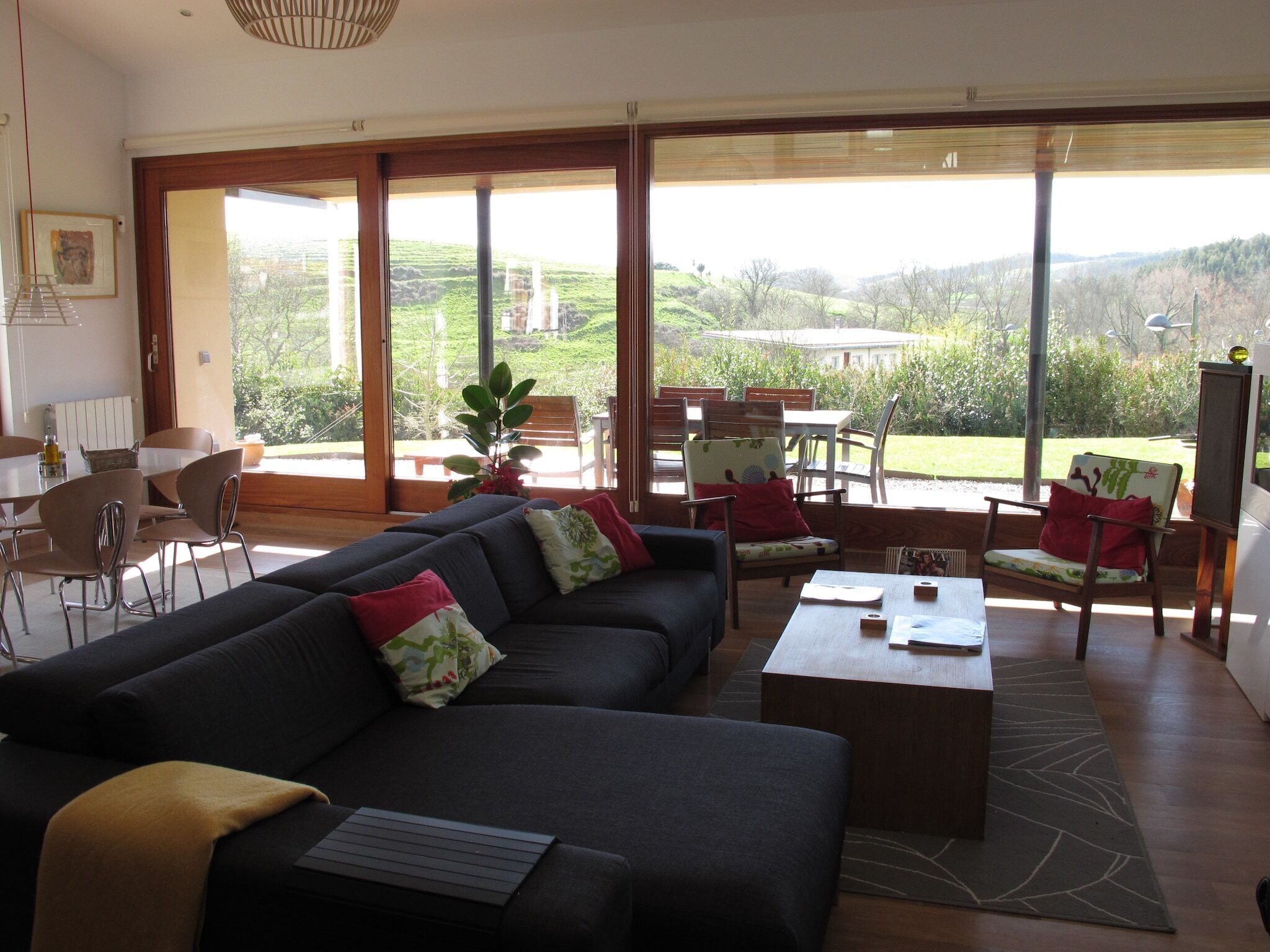 A spacious living area with a large sofa, coffee table, and chairs. There is a dining table with chairs, and a view of the outdoors through large windows.