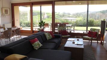 A spacious living area with a large sofa, coffee table, and chairs. There is a dining table with chairs, and a view of the outdoors through large windows.