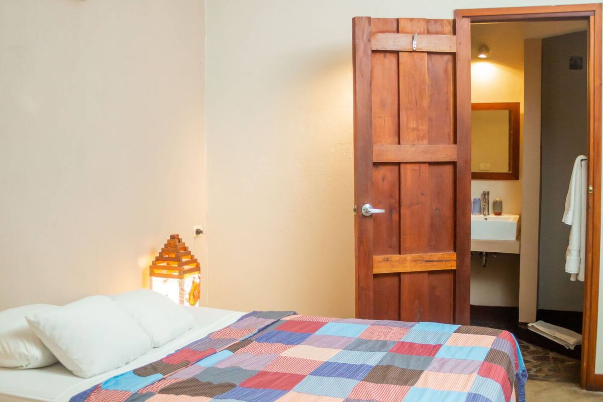 A bedroom with a bed, a wooden door, a bathroom with a sink, and a wall-mounted air conditioner.