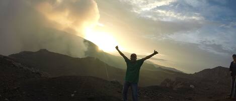 A person standing with arms raised in a rocky outdoor setting, with a mountain and a cloudy sky in the background.