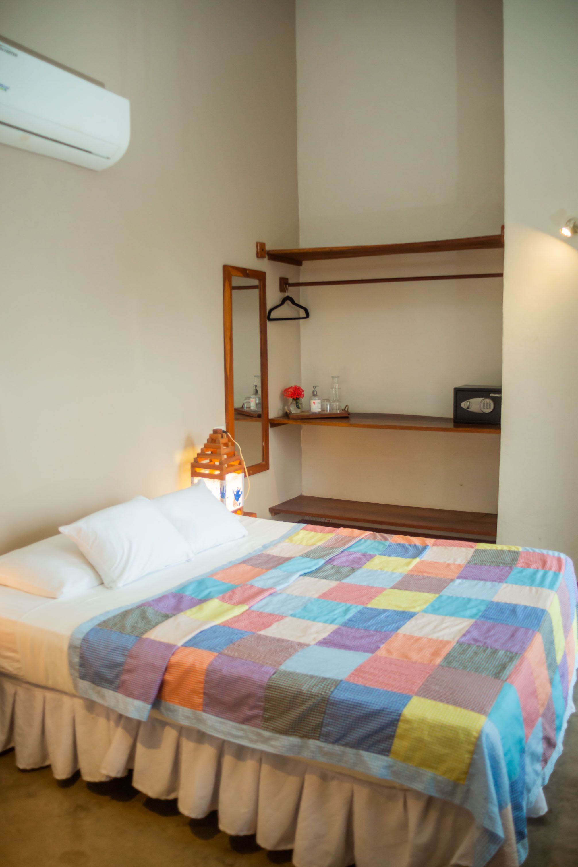 Un dormitorio con cama, colcha colorida, mesita de noche con lámpara, espejo y estante con objetos.