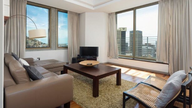 A modern living room with a sofa, a coffee table, and a TV. Large windows offer a view of the cityscape. - ITC Hospitality Group Two Bedrooms Cartwright's Corner (Cape Town)