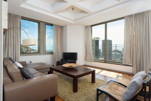Un salon moderne avec un canapé, une table basse et un téléviseur. De grandes fenêtres offrent une vue sur le paysage urbain.