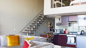 A modern living room with a grey sofa, yellow and red cushions, a staircase, and a kitchen area with a dishwasher and stove. - ITC Hospitality Group Two Bedrooms Cartwright's Corner (Cape Town)