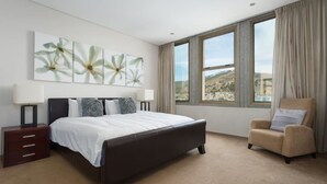 A bedroom with a large bed, a chair, a nightstand, and a window with a view of a landscape. - ITC Hospitality Group Two Bedrooms Cartwright's Corner (Cape Town)
