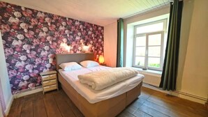 A bedroom with a floral wallpaper, a wooden floor, a bed with white bedding, a nightstand, and a window with curtains. - Holiday Home in Hotton near Caves of Hotton (Werpin)