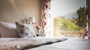 Una camera da letto con un letto, una lampada, un cuscino floreale, un vaso con fiori e vista su vegetazione e montagne attraverso una finestra.