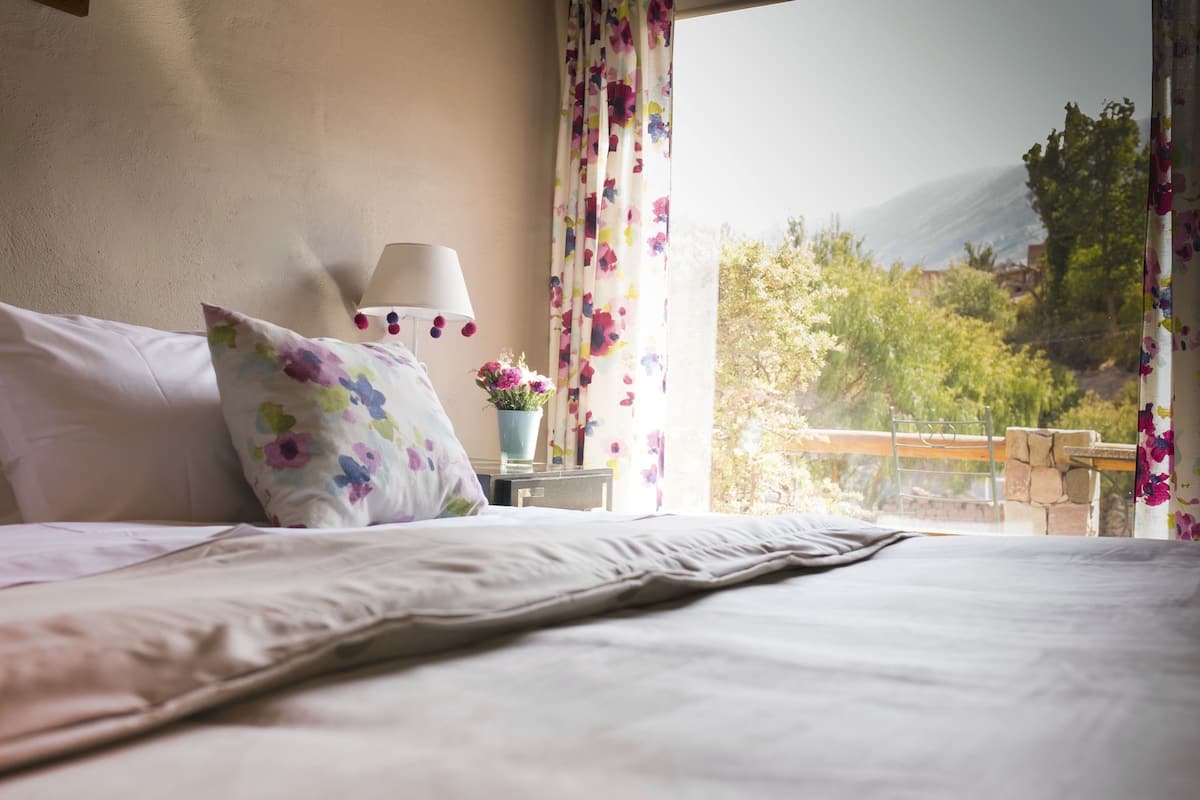A bedroom with a bed, a lamp, a floral pillow, a vase with flowers, and a view of greenery and mountains through a window.