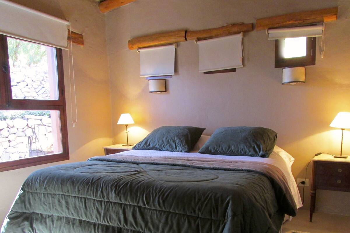 A bedroom with a large bed, two bedside lamps, and a window with a view of a stone wall.
