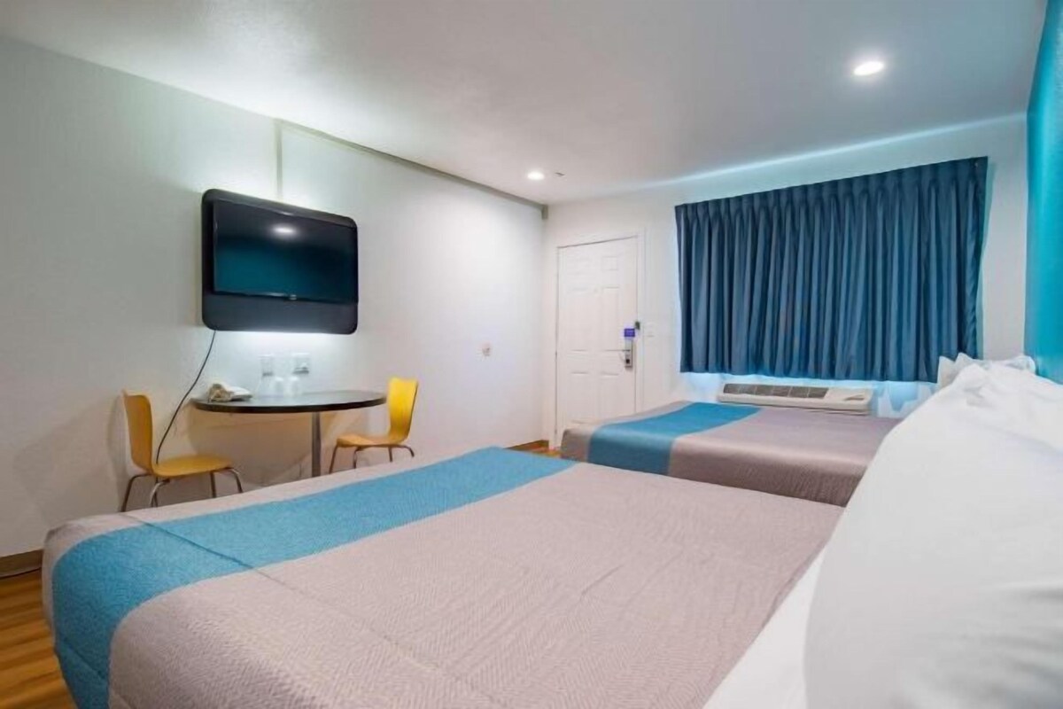 Quarto de hotel com duas camas, uma escrivaninha com cadeira, televisão e janela com cortinas.