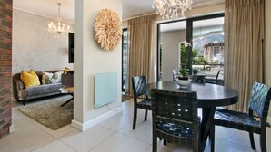 A modern living room with a sofa, a dining table with chairs, a chandelier, and a large window with curtains. - ITC Hospitality Group One Bedrooms Rockwell Building (Cape Town)