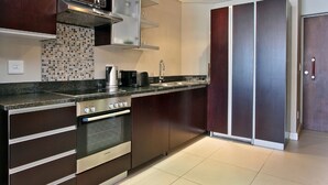 A modern kitchen with dark cabinetry, a stainless steel oven, a tiled backsplash, and a double sink. - ITC Hospitality Group One Bedrooms Rockwell Building (Cape Town)