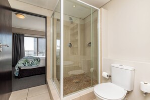 Een moderne badkamer met een glazen douchecabine, toilet en een bed met uitzicht op de stad.