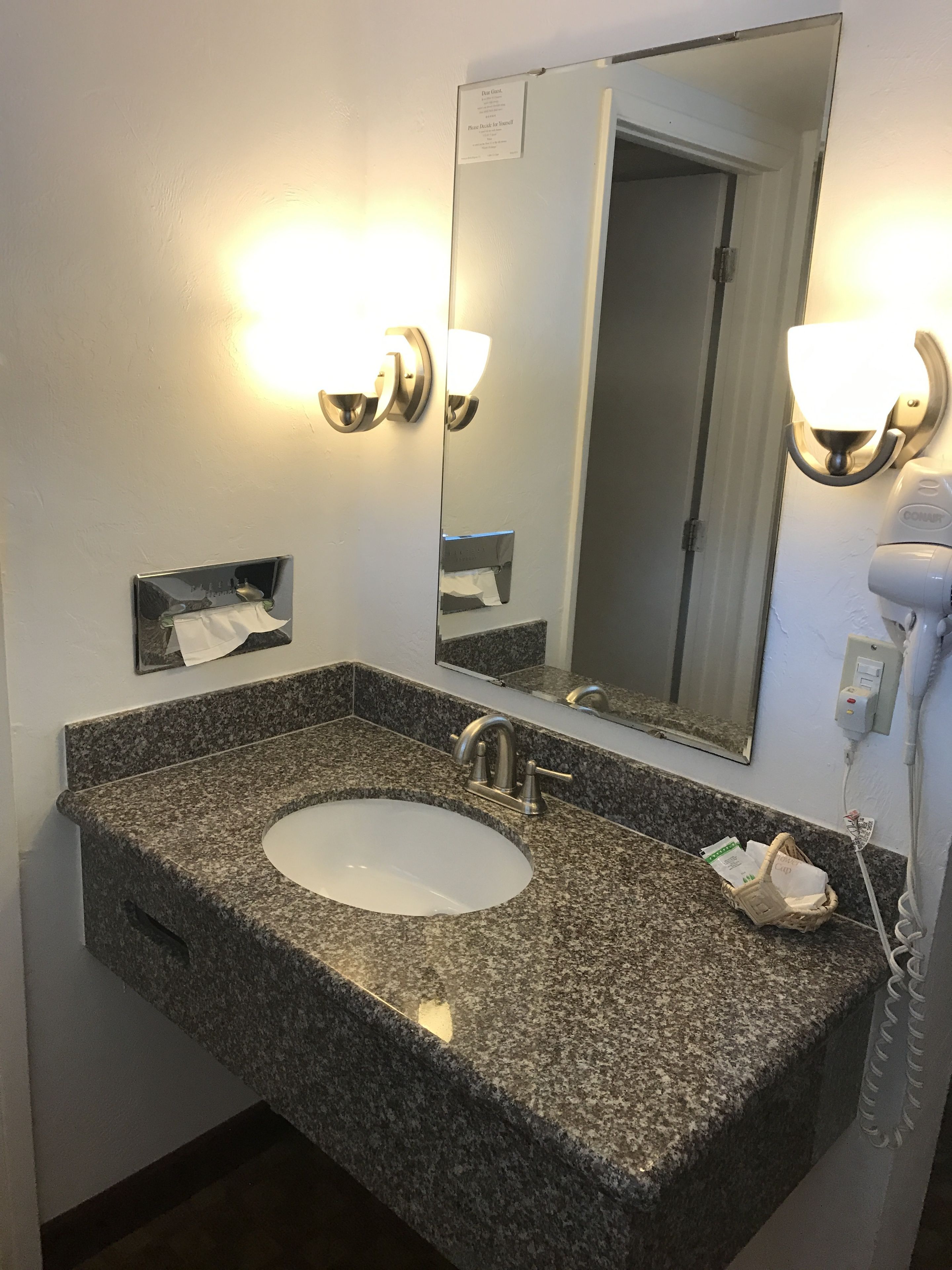 Un baño con lavamanos de granito, un espejo y un secador de pelo en la pared.