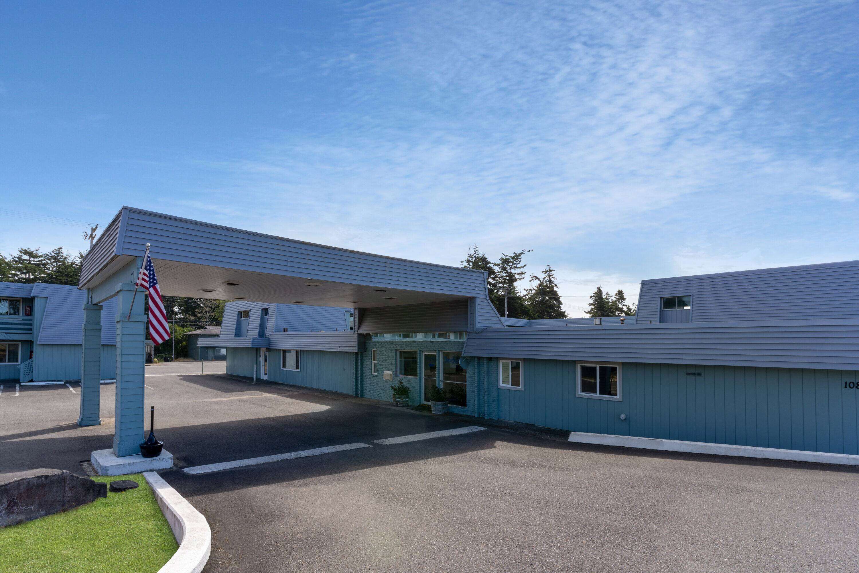 A motel with a covered parking area, a flagpole with an American flag, and a clear blue sky.