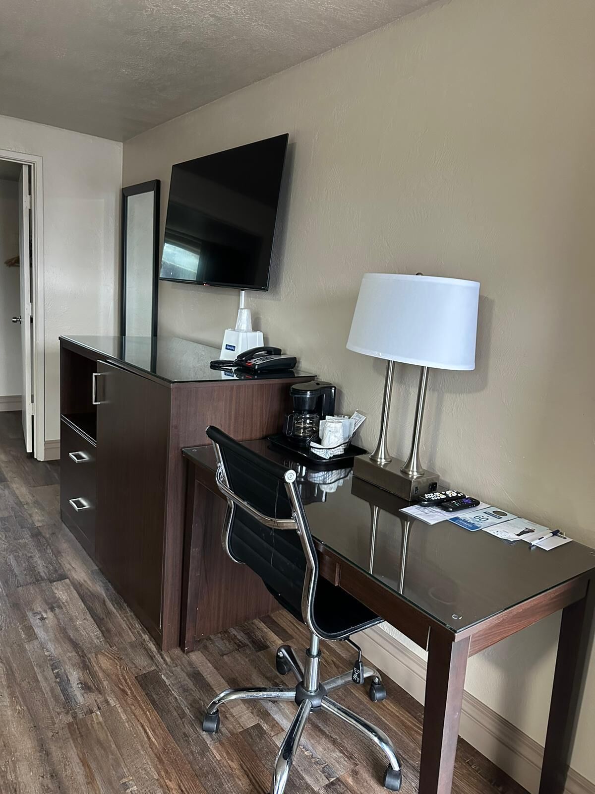 A hotel room with a wooden desk, a chair, a flat-screen TV, a lamp, a coffee maker, and a minibar.