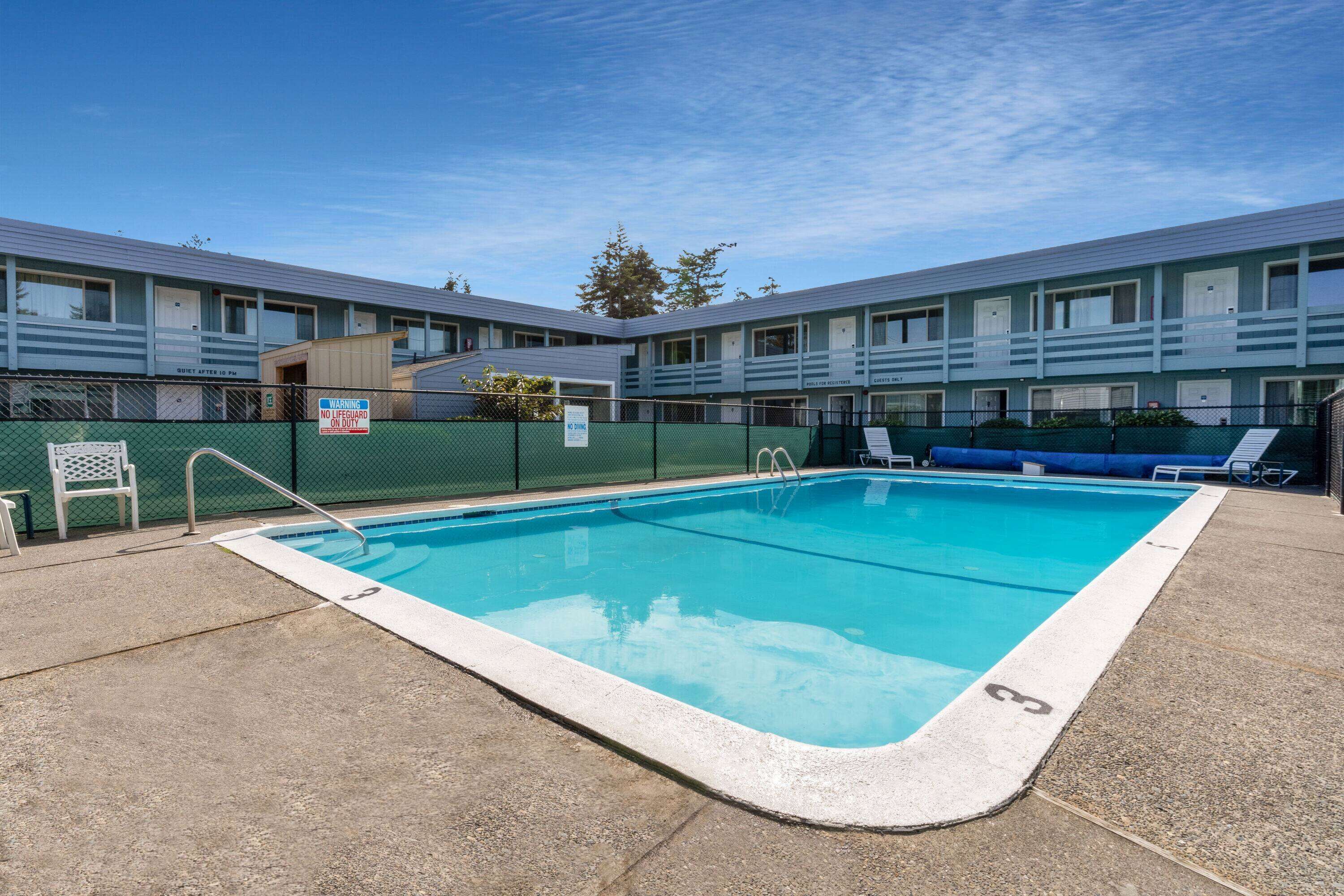 Una piscina con area recintata, un'area relax coperta con poltrone a sdraio e un edificio motel con diverse camere.