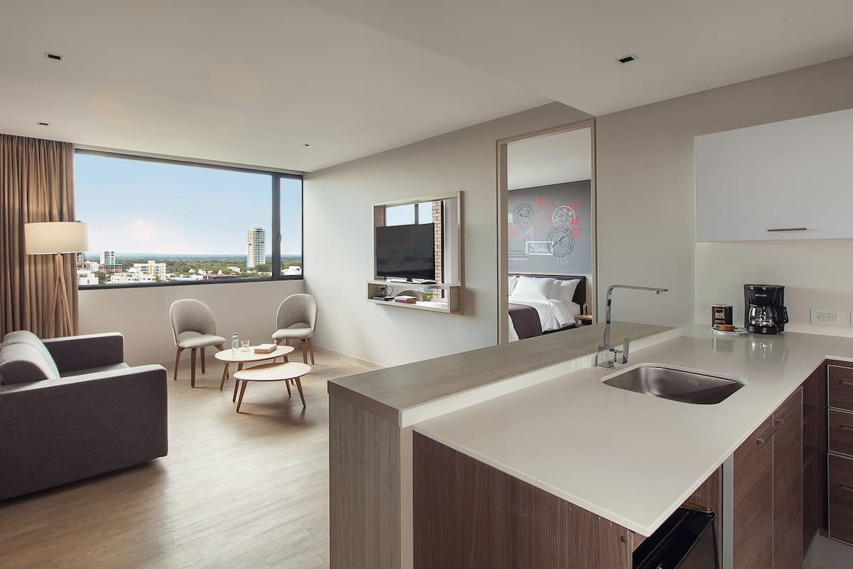 A modern hotel room with a sofa, chairs, a small dining table, a TV, a kitchen area with a sink, and a city view.