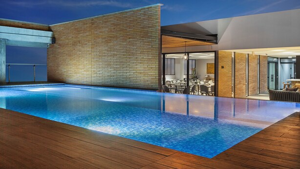 An infinity pool with a wooden deck, a brick wall, and a modern interior with glass doors and a dining area. - Hotel Sites Monteria (Montería)