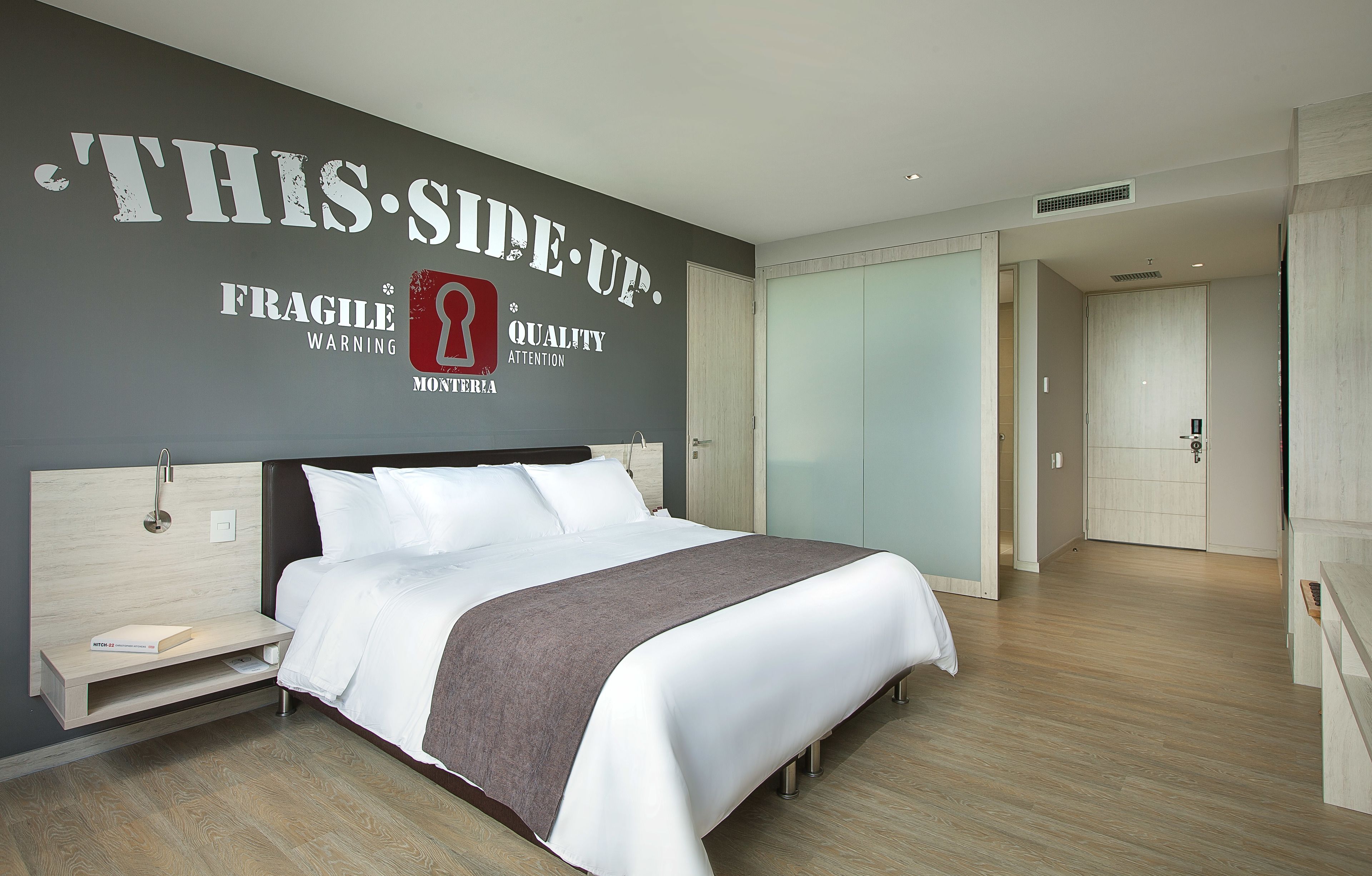 Quarto de hotel moderno com uma cama grande, uma mesa de cabeceira e um criado-mudo, e uma parede com sinal de segurança.