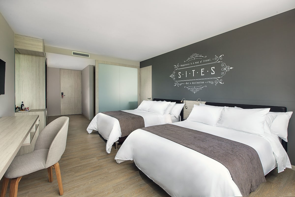 A hotel room with two beds, a desk, and a chair. There is a wall with decorative text and a vent.