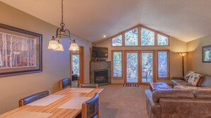 A spacious living room with a fireplace, a dining area, and a large window with a view of trees. - Riverwood on Fall River (Estes Park)