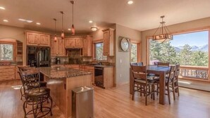 A spacious kitchen with wooden cabinets, a central island, bar stools, and a dining area with a view of mountains. - Riverwood on Fall River (Estes Park)
