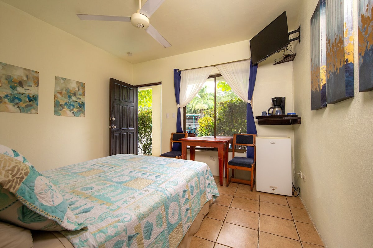 A bedroom with a bed, a desk, a chair, a refrigerator, and a ceiling fan.