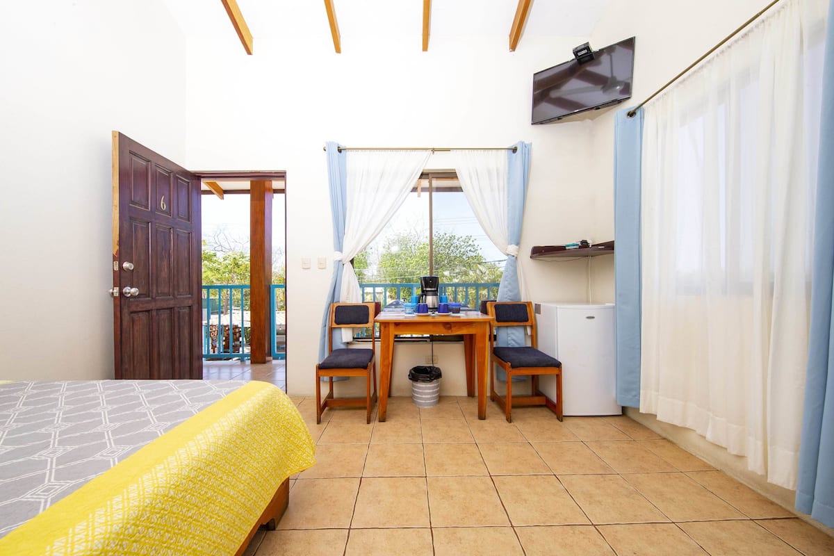 A room with a bed, a table with chairs, a small fridge, and a TV mounted on the wall.