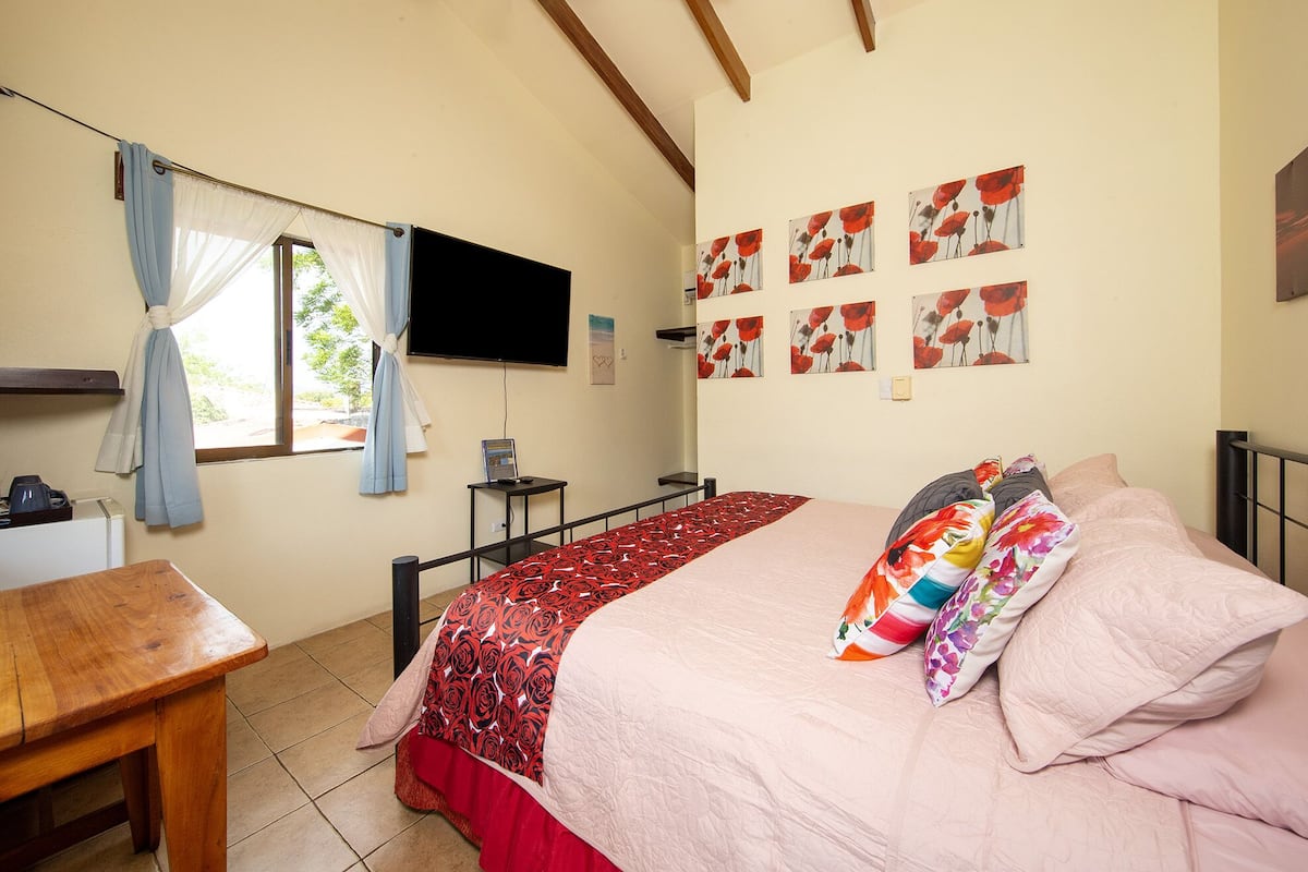A bedroom with a bed, a desk, a television, and a window with curtains.