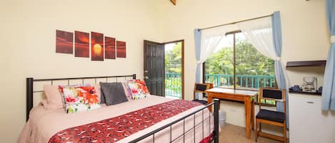 A bedroom with a bed, a desk, and a chair. There is a window with curtains, a door, and a view of greenery outside.