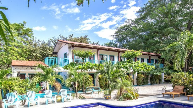 Una piscina con borde de baldosas, rodeada de palmeras y un edificio con techo rojo.