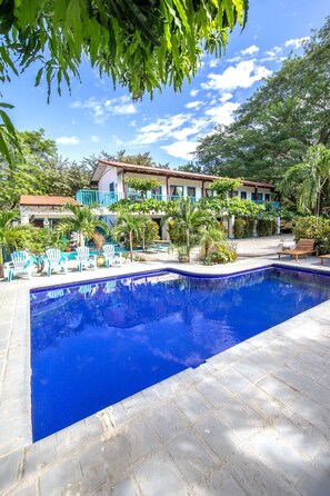 A swimming pool with a tiled deck, surrounded by palm trees and a building with a red roof. - Hotel Diversion Tropical (Cabo Velas)