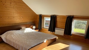 Une chambre avec un plafond en pente en bois, un grand lit, une table de chevet, une fenêtre avec des rideaux et une vue sur un paysage verdoyant.