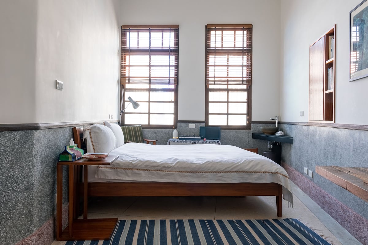 A bedroom with a wooden bed, two windows with blinds, a bedside table, a chair, a desk, a mirror, and a small shelf.