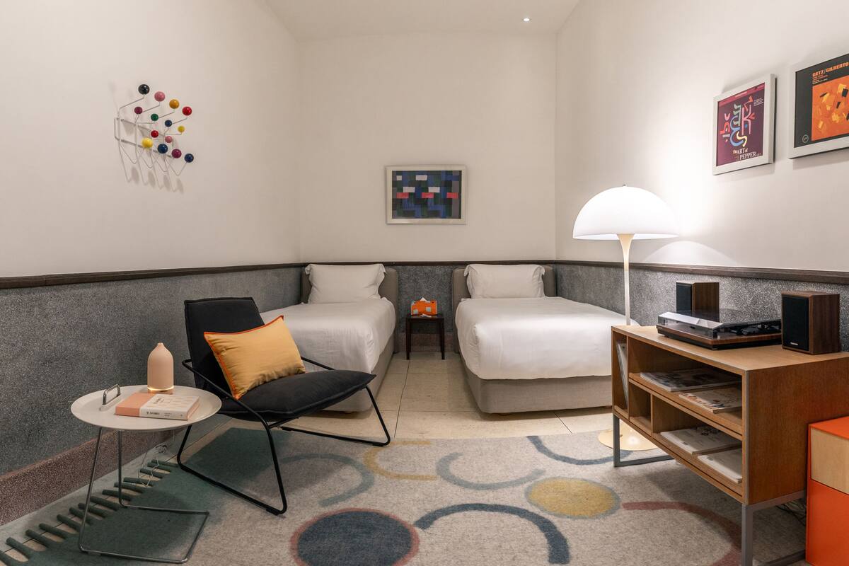 A hotel room with two beds, a chair, a small table, a lamp, and a shelf with books and a speaker.