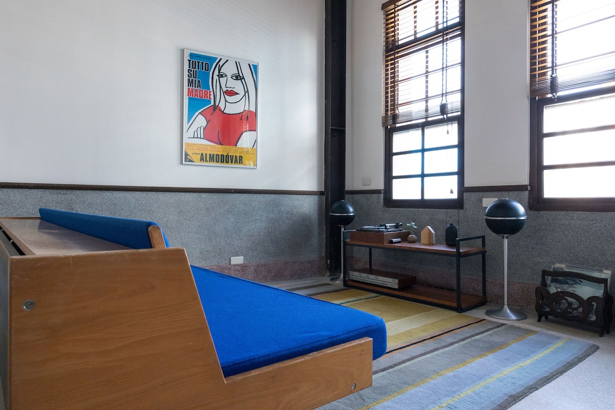 A room with a wooden bed, a small table with a lamp, a poster on the wall, and a window with blinds.