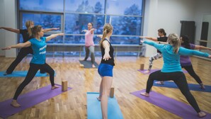 Un groupe de personnes participe à un cours de yoga, debout sur des tapis dans une pièce dotée de grandes fenêtres et d’un plancher en bois.