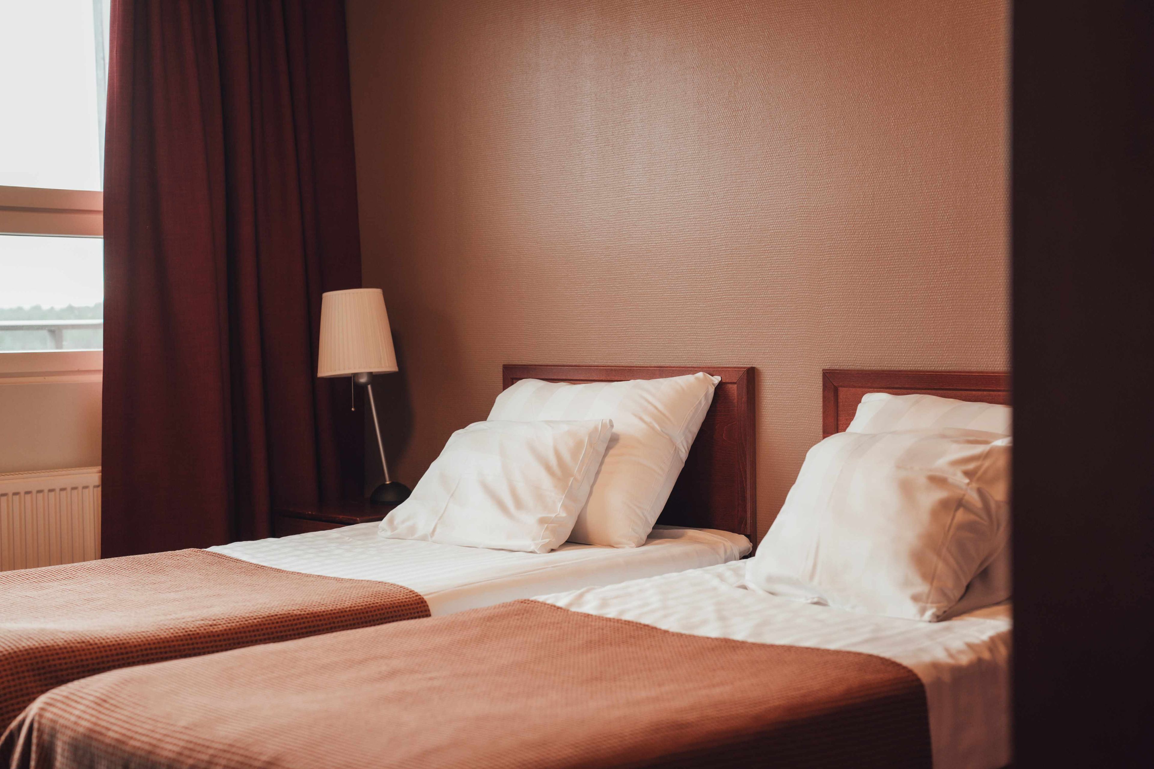 A hotel room with two single beds, a nightstand with a lamp, and a window with curtains.
