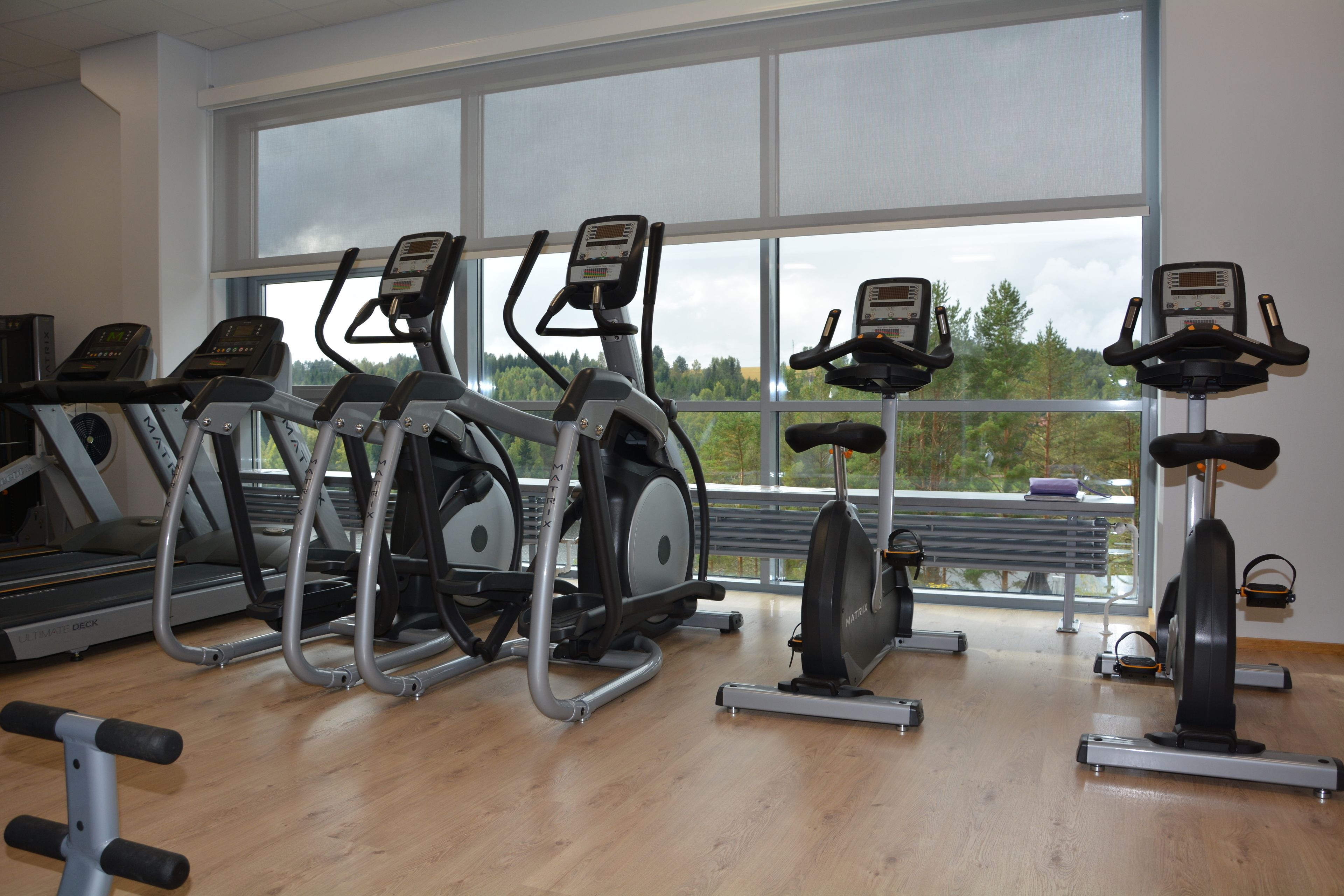 Un gimnasio con diferentes máquinas de cardio, ventanas amplias y suelo de madera.