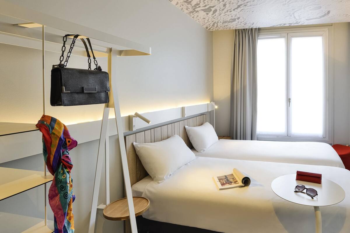 Una habitación de hotel con una cama, una mesita de noche con vasos, un bolso colgado y una bufanda colorida.