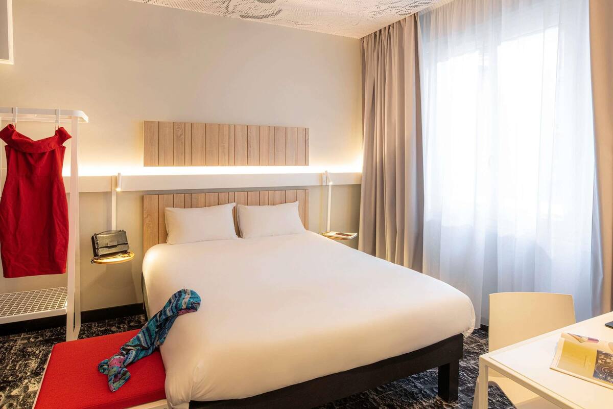 Habitación de hotel con una cama grande, un escritorio, una silla, un vestido rojo colgado y una maleta en un estante.