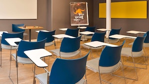 Une salle de classe avec des chaises bleues et des tables blanches, un écran de projection et une affiche accrochée au mur.