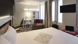 Une chambre d’hôtel avec un grand lit, un bureau avec une chaise, une petite table avec une lampe, une carte sur le lit et un téléviseur fixé au mur.
