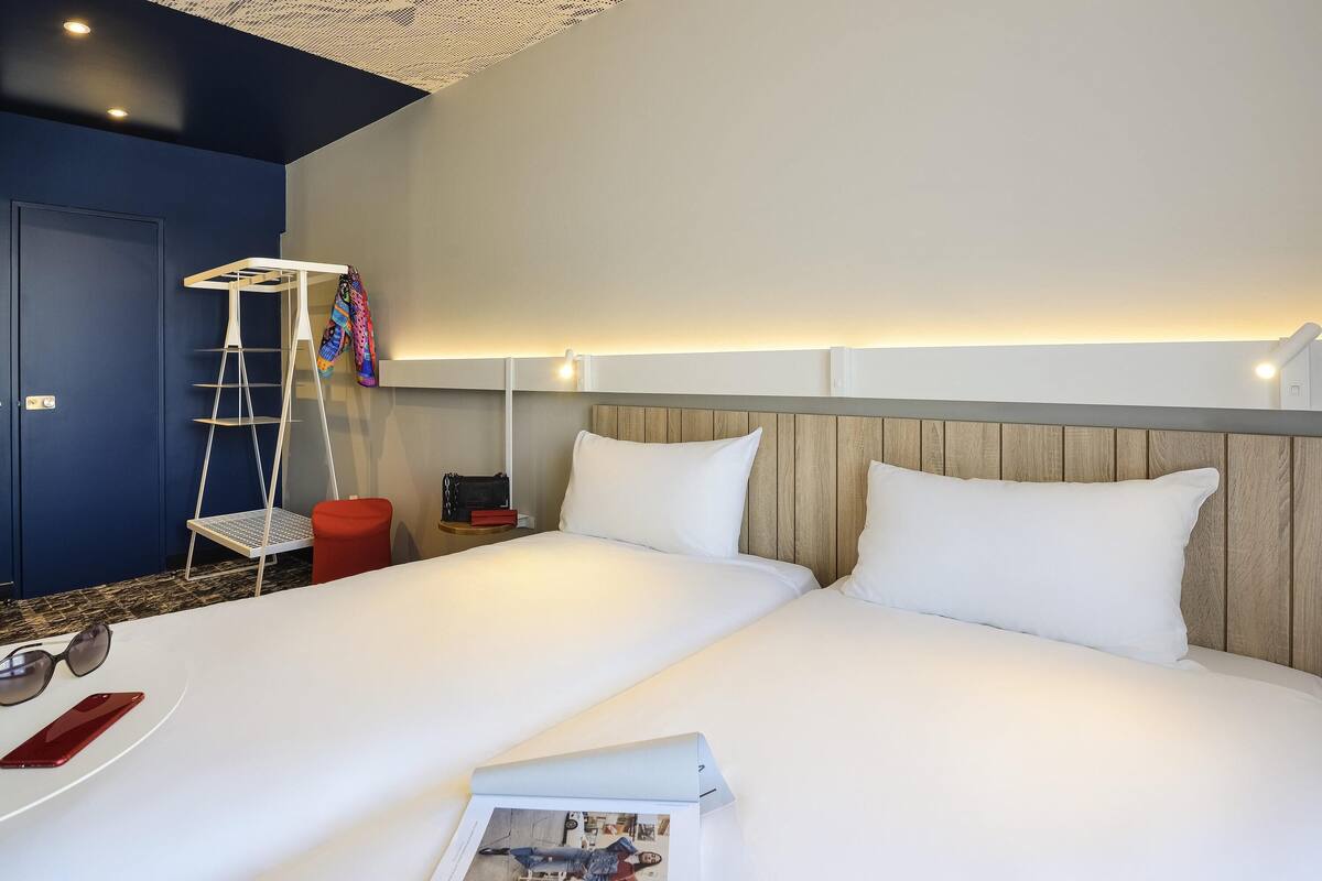 Una habitación de hotel con una cama grande, una mesita de noche, una silla roja, una estantería escalonada y una lámpara de pared.